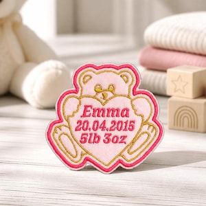 May include: A pink and gold embroidered patch shaped like a teddy bear holding a heart. The heart features the embroidered text "Emma", "20.04.2015", and "5lb 3oz" in dark pink thread, with a gold border outlining the bear and heart shapes.