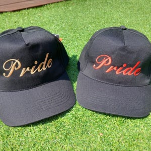 May include: Two black baseball caps with the word "Pride" embroidered on the front. One cap has gold embroidery and the other has red embroidery.