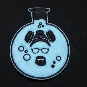 May include: A blue felt patch with an embroidered image of a person with glasses and a beard inside a beaker. The beaker has a black rim and a black stopper. There are small circles around the person's head.