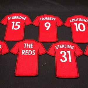May include: Seven red fabric patches shaped like football shirts, each featuring white text and numbers. The names include STURRIDGE 15, LAMBERT 9, COUTINHO 10, LALLANA 20, THE REDS, STERLING 31, and BALOTELLI 45, all displayed in a clear, bold font.