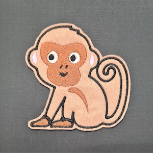 May include: An embroidered brown monkey with a curled tail, pink ears, and black eyes. The monkey is sitting on a grey background.