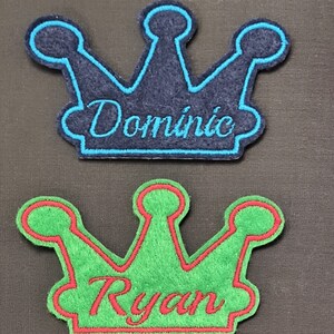 May include: Two felt crown patches with embroidered names. The top patch is navy blue with a turquoise outline and the name "Dominic" embroidered in turquoise. The bottom patch is green with a red outline and the name "Ryan" embroidered in red.