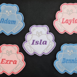 May include: Five embroidered felt patches shaped like teddy bears holding hearts. Each patch has a different name embroidered on the heart: Adam, Layla, Isla, Ezra, and Dennis.