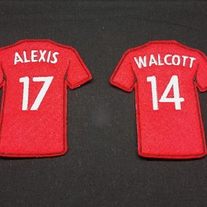 May include: Two red felt patches shaped like sports jerseys. The left patch features the white text ALEXIS above the number 17. The right patch features the white text WALCOTT above the number 14. Both patches have dark stitched outlines.