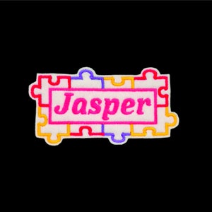 May include: A white fabric patch with a colourful puzzle piece border and the name "Jasper" in pink lettering.