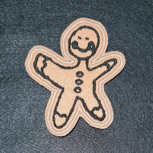 May include: A tan gingerbread person patch with black embroidered outlines and facial features. The design includes three small black circles down the center of the torso and black accents on the hands and feet, all set against a dark gray fabric.