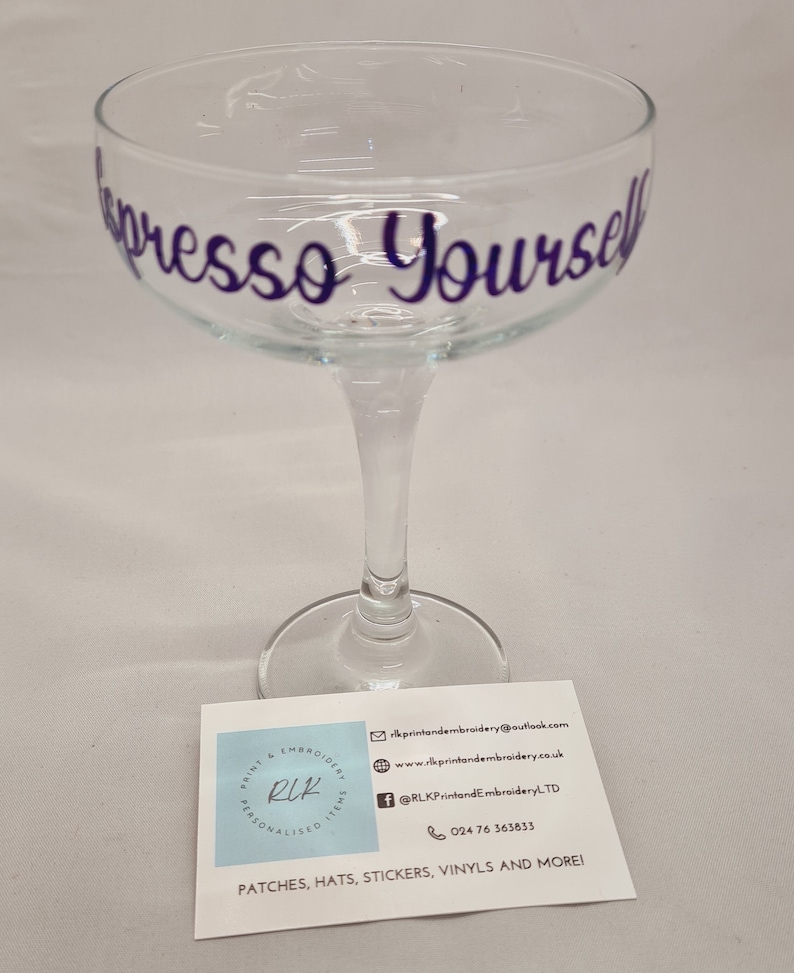 Puede incluir: Una copa de champ&aacute;n de cristal transparente con el texto "Espresso Yourself" escrito en morado en el lateral. La copa est&aacute; sobre una superficie blanca junto a una tarjeta de visita con la informaci&oacute;n de contacto de una empresa que ofrece art&iacute;culos personalizados.