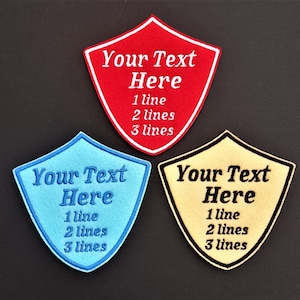 May include: Three embroidered iron-on patches in the shape of shields. The patches are red, blue, and yellow. Each patch has the text "Your Text Here" and the text "1 line, 2 lines, 3 lines".
