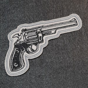 May include: A black and white embroidered patch of a revolver pistol with a silver outline.