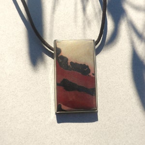 May include: A rectangular pendant featuring a smooth, multi-colored surface with bands of cream, deep red, and black, encased in a thin silver-toned metal frame. The pendant is suspended from a simple, dark brown cord necklace.