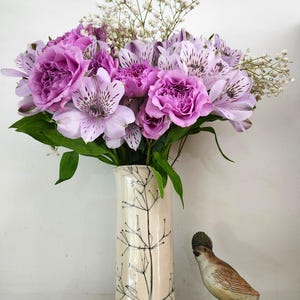 May include: A tall, cylindrical, cream-colored ceramic vase decorated with a black line drawing of a plant stem. The vase holds a vibrant bouquet of purple roses, light purple alstroemeria with dark speckles, and white baby's breath flowers.