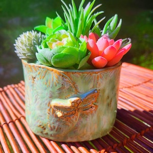 May include: A cylindrical ceramic planter with a light green glaze and a textured dragonfly relief on the front. The planter is filled with an arrangement of various artificial succulents in shades of bright green, muted sage, and vibrant pinkish-red.