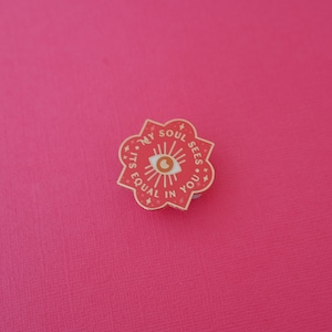 Puede incluir: Un pin de esmalte rojo con un contorno dorado en forma de flor con un ojo estilizado en el centro. El texto alrededor del ojo dice "My Soul Sees Its Equal In You."
