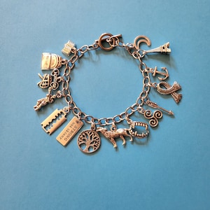 May include: Silver charm bracelet with various charms, including a tree, wolf, razor blades, and the Eiffel Tower. The bracelet has a toggle clasp and is displayed on a blue background. The charms are detailed and add a unique touch to the jewelry.