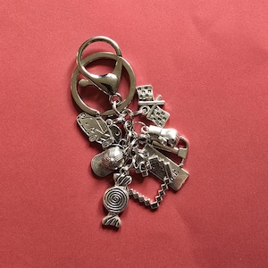 May include: Silver-toned keychain with multiple charms, including a car, a baseball cap, a candy, a house, a boxing glove, a heart, and a sign. The keychain has a lobster clasp and a split ring. The charms are detailed and have an antique finish.