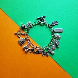 May include: A silver charm bracelet with a variety of charms, including a backpack, binoculars, a dice, a compass, a basketball, a bicycle, and a charm that says "NERD". The bracelet also has a charm that says "BRAVE".