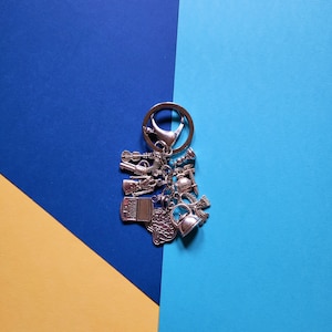 May include: Silver-toned keychain with multiple charms, including a laptop, teapot, and tools. The keychain is on a background of blue and yellow geometric shapes. The charms are detailed and appear to be made of metal.