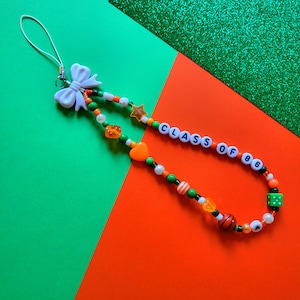 May include: A white bow charm with a green, orange, and gold beaded phone charm with the text "CLASS OF 86".