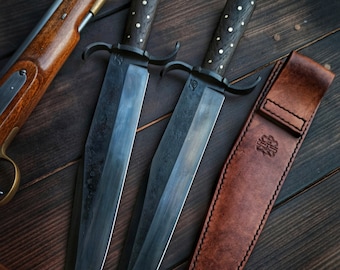 May include: Two black-handled knives with ornate metal guards and leather sheaths. The blades are long and sharp, with a slight curve. The handles are decorated with small brass dots. The knives are laid out on a dark wooden surface.