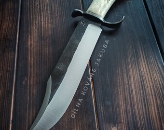 May include: A black-handled knife with a white antler handle. The blade is made of steel and has a sharp point. The knife is on a wooden surface. The text "DILNA KOVARNE JAKUBA" is engraved on the blade.