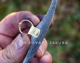 May include: A hand-forged metal tool with a curved blade and a brass ring. The tool has a textured, dark gray finish and a brass accent. The text "DILNA KOVÁŘE JAKUBA" is visible. The tool's design suggests it may be used for leatherworking or similar crafts.
