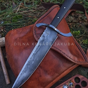 Könnte beinhalten: Ein großes, handgefertigtes Messer mit einem dunklen Holzgriff und einer gezahnten Klinge. Das Messer hat einen Parier und wird auf einer Ledertasche präsentiert. Der Text "DÍLNA KOMÁŘE JAKUBA" ist auf der Tasche zu sehen.
