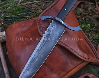May include: A large, handmade knife with a dark wooden handle and a serrated blade. The knife features a crossguard and is displayed on a leather satchel. The text "DÍLNA KOMÁŘE JAKUBA" is visible on the satchel.