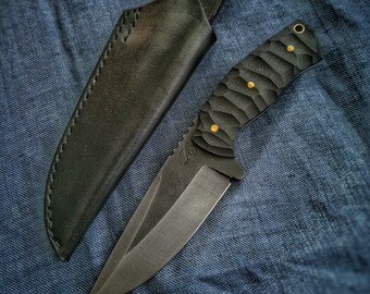 May include: A black knife with a textured black handle and a leather sheath. The knife has a gold inscription that reads "DÍLNA KOVAŘE JAKUBA".
