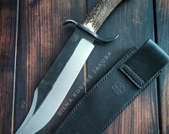 May include: A large hunting knife with a polished steel blade and a textured antler handle. The knife rests on a dark wooden surface next to a black leather sheath. The blade has a curved edge and a small logo. The sheath has a logo and the text "DÍLNA KOVÁŘE JAKUBA".