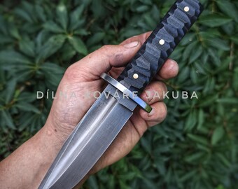 May include: A handcrafted knife with a dark, textured handle and a polished silver blade. The handle features three gold-colored rivets. The text "DÍLNA KOVÁŘE JAKUBA" is visible. The knife is held against a backdrop of green foliage.