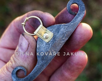 May include: A hand-forged metal keychain with a unique, curved design. The keychain features a brass ring and a rectangular brass plate riveted to the metal. The metal has a textured, dark gray appearance. The text "DILNA KOVÁŘE JAKUBA" is visible.
