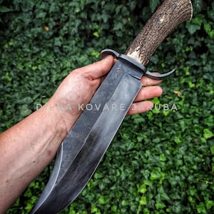 Puede incluir: Un cuchillo grande, artesanal, con una hoja oscura y texturizada y un mango de asta. El cuchillo tiene una guarda y una construcción de espiga completa. El mango es de color marrón claro con una punta blanca. El fondo es hiedra verde.
