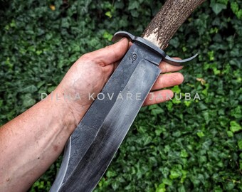 May include: A large, handcrafted knife with a dark, textured blade and a handle made from antler. The knife has a guard and a full tang construction. The handle is a light brown color with a white tip. The background is a green ivy.