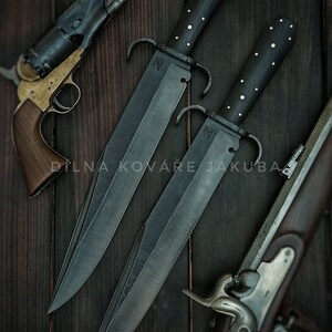 Könnte beinhalten: Zwei Messer mit schwarzen Griffen, verzierten Details und einer silbernen Inschrift auf der Klinge. Die Messer liegen auf einer dunklen Holzoberfläche mit einer Schwarzpulverpistole und einem Steinschlossgewehr.