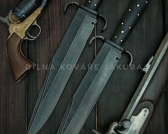 May include: Two black-handled knives with ornate details and a silver inscription on the blade. The knives are laid out on a dark wood surface with a black powder pistol and a flintlock rifle.