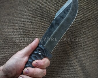 May include: A black knife with a textured handle and a sharp blade. The knife is held in a hand and the text "DÍLNA KOVÁŘE JAKUBA" is visible on the background.