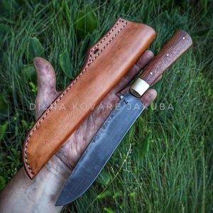 Op de afbeelding: Een handgemaakt mes met een donker lemmet en een houten handvat met messing accenten. Het mes wordt getoond met een bruine leren schede. De tekst "DILNA KOVÁŘE JAKUBA" is zichtbaar.