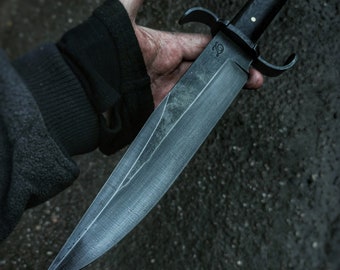 May include: A black-handled knife with a long, sharp blade. The blade has a textured finish and a slight curve near the handle. The handle has a black wood finish and a metal guard with two prongs.