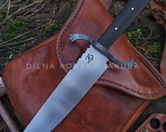 May include: A large, handmade knife with a dark wooden handle and a serrated blade. The blade has a decorative emblem and the text "DÍLNA KOVÁŘE JAKUBA". The knife rests on a brown leather bag.