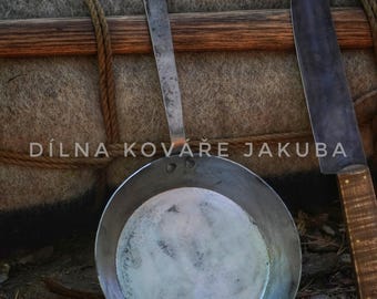 May include: A handmade, rustic metal frying pan with a long handle and a sharp knife with a wooden handle. The text "DÍLNA KOVÁŘE JAKUBA" is visible. The items are set against a backdrop of a gray, textured surface.