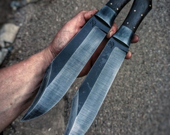 May include: Two black-handled knives with a dark gray finish and a slight curve to the blade. The knives are held in a hand with the blades pointing towards the camera. The text "DÍLNA KOVAŘE JAKUBA" is visible at the bottom of the image.