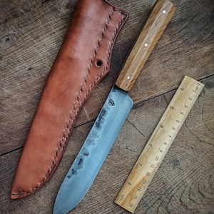 Op de afbeelding: Een handgemaakt mes met een bruin houten handvat en een leren schede. Het blad is van staal en heeft een gehammerde afwerking. Het mes is 20 cm lang.