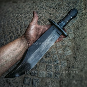 May include: A large, black, hand-forged knife with a wooden handle. The blade is curved and has a sharp point. The handle is made of dark wood with a textured surface. The knife is held in a hand with dirty fingers. The text "DÍLNA KOVÁŘE JAKUBA" is visible on the background.