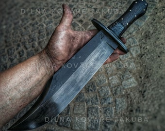 May include: A large, black, hand-forged knife with a wooden handle. The blade is curved and has a sharp point. The handle is made of dark wood with a textured surface. The knife is held in a hand with dirty fingers. The text "DÍLNA KOVÁŘE JAKUBA" is visible on the background.