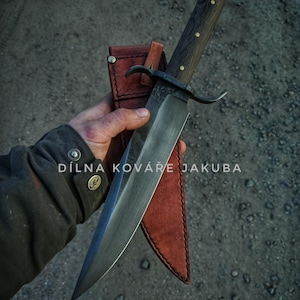 Op de afbeelding: Een groot, zwart, handgesmeed mes met een houten handvat en een leren schede. Het mes heeft een gebogen lemmet en een decoratief ontwerp op het handvat. De tekst "DÍLNA KOVÁŘE JAKUBA" is zichtbaar op het lemmet.