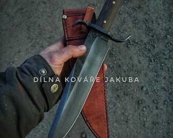 May include: A large, black, hand-forged knife with a wooden handle and a leather sheath. The knife has a curved blade and a decorative design on the handle. The text "DÍLNA KOVÁŘE JAKUBA" is visible on the blade.