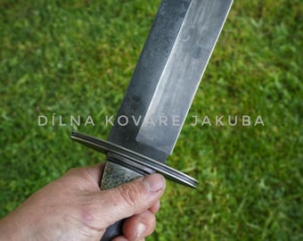 May include: A large, handcrafted knife with a dark, textured blade and a black wooden handle. The knife has a crossguard and is held against a green grassy background. The text "DÍLNA KOVÁŘE JAKUBA" is visible.