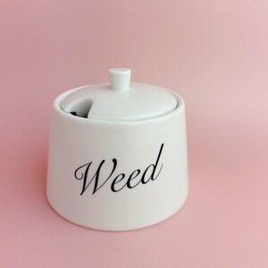 Może przedstawiać: Biały ceramiczny słoik z dopasowaną pokrywką z małym uchwytem i wycięciem na łyżeczkę. Słowo Weed jest nadrukowane czarną kursywą na przedniej części słoika, który ma zwężający się cylindryczny kształt oraz gładkie wykończenie powierzchni.