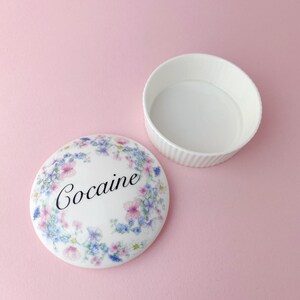May include: A white ceramic trinket box with a floral design and the word "Cocaine" written in black lettering. The lid is off and the box is sitting on a pink surface.