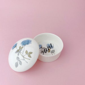 May include: A white ceramic trinket box with a lid, decorated with blue floral patterns. The open box reveals the word "bunt". The background is a pale pink.